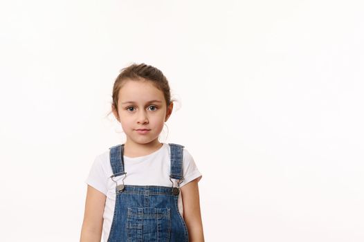 Isolated portrait on white background of a Caucasian adorable blue-eyed child, cute baby girl in white t-shirt and blue denim overalls, confidently looking at camera. Copy space for advertising text