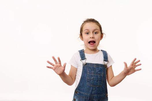 Emotional portrait on white background of a preschool child, overjoyed cute baby girl, wearing blue denim overalls, sweetly smiling, showing her hands palms to camera. Beautiful kids, happy childhood