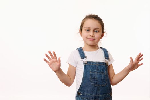 Emotional portrait on white background of beautiful preschool child, cute baby girl, wearing blue denim overalls, sweetly smiling, showing her hands palms to camera