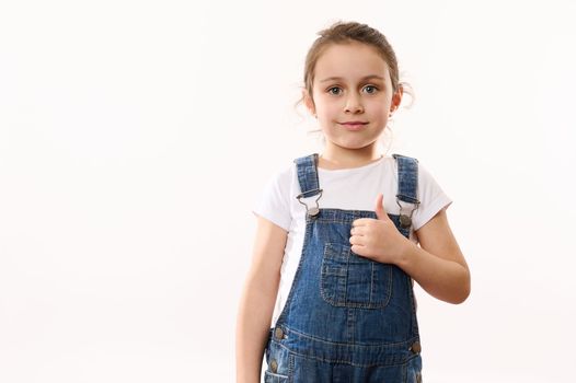 Beautiful European elementary age, preschool, first grader little child girl, wearing blue denim overalls, shows a thumb up, cutely smiles looking at camera, isolated over white background. Copy space