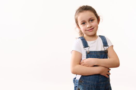Happy cute adorable little child baby girl in blue denim overalls and white t-shirt, hugging herself, smiling toothy smile looking at camera, isolated over white background with copy advertising space