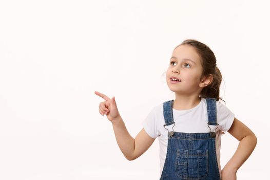 Advertising studio shot of an adorable Caucasian little child, cute baby girl in blue denim overalls, pointing her finger and looking at a copy ad space for promo text, isolated over white background
