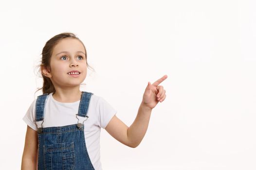 Horizontal studio shot on white background of a happy European little girl, adorable preschooler smart child, wearing blue denim overalls, pointing away on free space for your advertising content