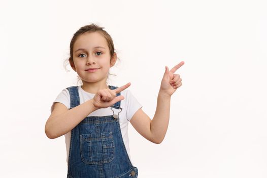 Isolated portrait on white background of a preschool Caucasian lovely child, adorable baby girl in blue denim overalls, pointing her finger at copy ad space, cutely smiling looking at the camera