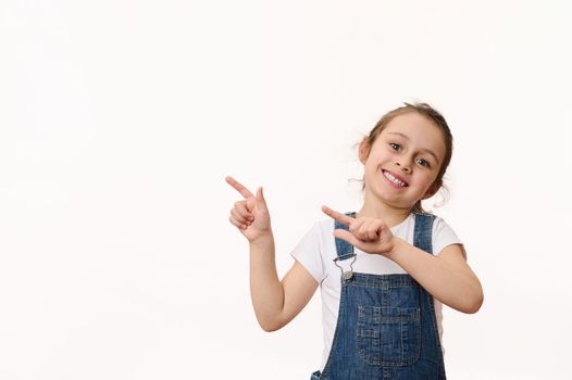 Adorable delightful Caucasian little child, a cute baby girl wearing a blue denim overalls, smiling a toothy smile, pointing at a copy space for promotional text, isolated over white background.