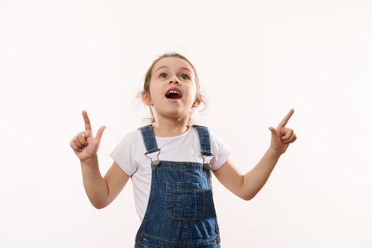 Beautiful overjoyed baby girl, astonished Caucasian little child wearing blue denim overalls, looks up and points with her finger at a copy space for promotional text, isolated over white background