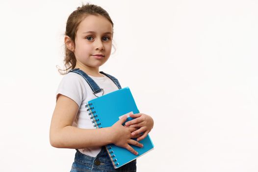 Adorable little girl in white t-shirt and blue casual denim, holding a copybook and cutely smiles looking at camera. Isolated portrait on white background of a smart cute preschooler child. Copy space