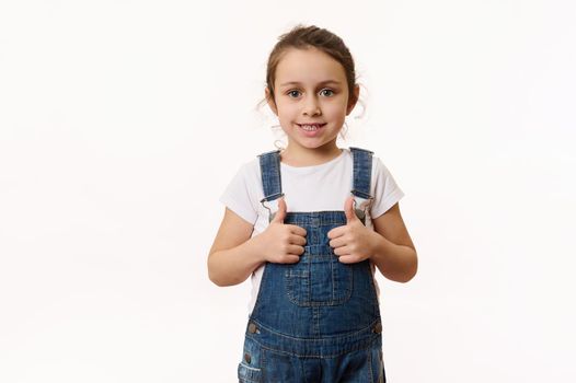 Adorable elementary age Caucasian little child, preschool kid, a lovely baby girl wearing blue denim overalls, shows thumbs up and sweetly smiles looking at camera, isolated on white background