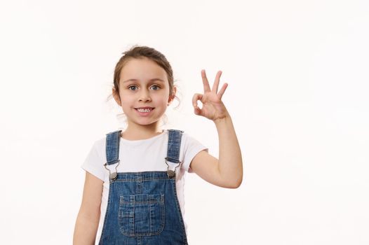Adorable elementary age, preschool little child girl, in blue denim overalls, gestures, shows peace sign, cutely smiling cheerful toothy smile, looking at camera, isolated white background. Copy space