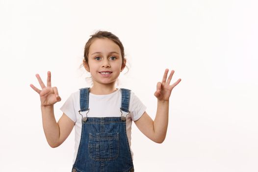 Adorable Caucasian little child, preschool kid, lovely baby girl wearing blue denim overalls, gestures, shows OK signs and happily smiles a toothy smile looking at camera, isolated on white background