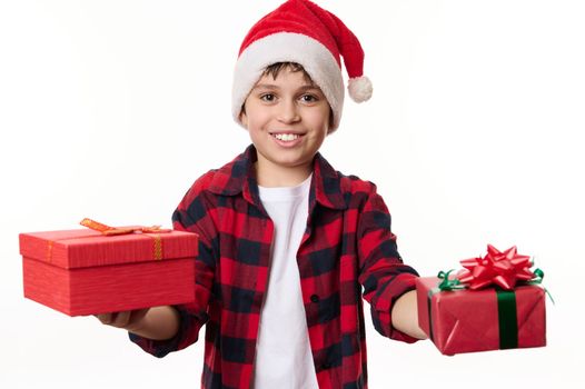 Handsome adorable multi-ethnic child boy in Santa hat, smiling a cheerful toothy smile, handing happy Christmas or New Year presents, looking at camera. Winter holidays. Magical Xmas mood. Pure joy