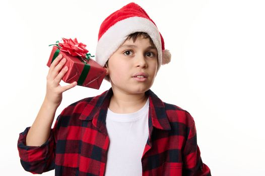 Curious child boy in Santa hat, shakes Christmas gift box, guessing the present inside it, isolated close-up portrait on white background with copy space. Time to open presents. Happy winter holidays