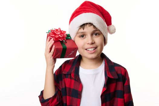 Adorable kid boy in Santa hat, shakes Christmas gift box, guessing the present inside it, smiling a beautiful smile looking at camera, isolated close-up portrait on white background with copy space