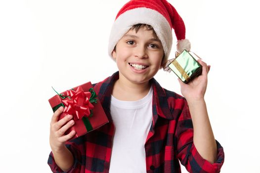 Adorable Hispanic boy wearing Santa hat, smiling a beautiful toothy smile, posing with Christmas gifts, looking at camera. New Years preparations. Winter holidays, magical festive atmospheric mood