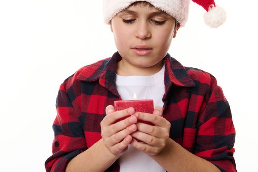 Close-up portrait of a handsome preteen Caucasian boy with a lit candle, isolated on white background. Adorable child making wish. Merry Christmas, Happy New Year and winter holidays. Copy ad space