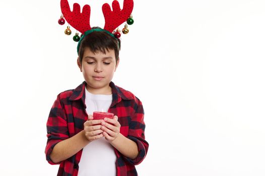 Half-length portrait of a boy in deer antler hoop, holding a red lit candle, making wish for Christmas, isolated on white background with copy advertising space. Magical atmosphere of winter holidays