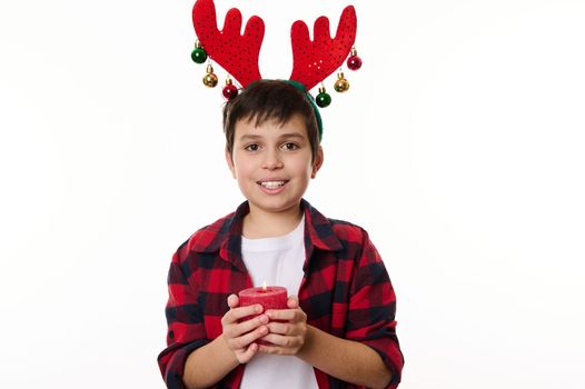 Happy Caucasian teenage boy with deer antler hoop on his head, smiling a beautiful toothy smile, holding a lit Christmas candle, looking at camera. Isolated on white background. Copy advertising space
