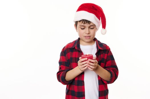 Handsome preteen boy in red checkered shirt and Santa hat, holding a decorative lit candle, making cherished wish for Christmas, isolated on white background. Happy childhood. Magical winter holidays