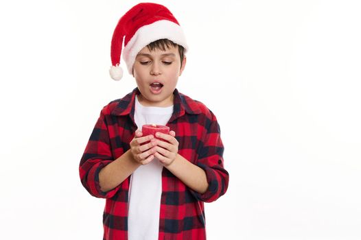 Isolated portrait on white background of an adorable Caucasian child boy, wearing Santa hat, making cherished wish for Christmas and blowing out red candle in his hands, Copy advertising space or text