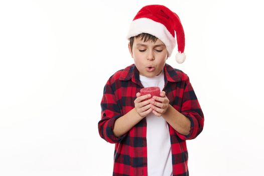 Caucasian preteen boy in red checkered shirt and Santa hat, makes cherished wish for Christmas, blows out a candle, isolated on white background. Happy childhood. Magical atmosphere at winter holidays