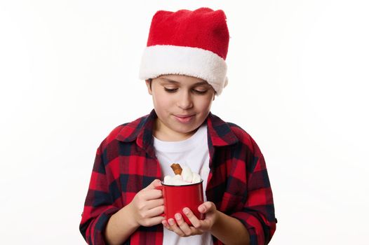 Adorable preteen boy wearing Santa hat, warming his hands with a red cup of hot chocolat and marshmallows, isolated on white background with copy advertising space. Merry Christmas, New Year concept