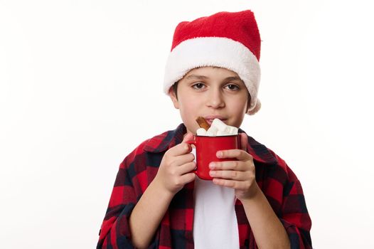 Isolated portrait on white background of a happy multi-ethnic child boy in Santa hat, enjoying a delicious sweet drink of hot cocoa with marshmallows. Christmas. Winter holidays concept. Copy ad space