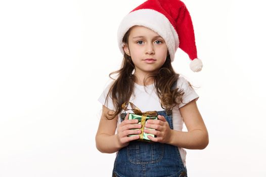Lovely baby girl, adorable little child wearing Santa hat and blue denim overalls, holds a happy Christmas present wrapped in green shiny gift paper, isolated on white. Christmas time. Winter holidays