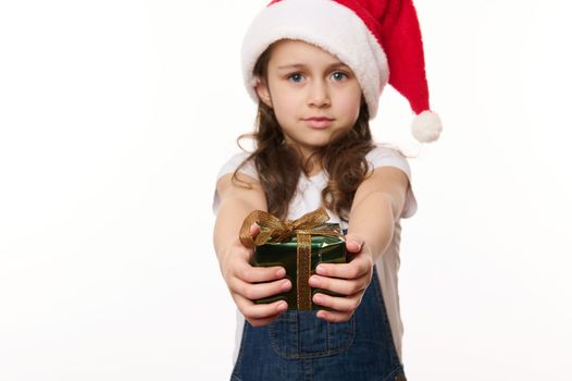 Caucasian cute baby girl, a little child wearing Santa hat and blue denim overalls, handing a happy Christmas present, looking at camera, isolated on white. Christmas time and New Year's preparations