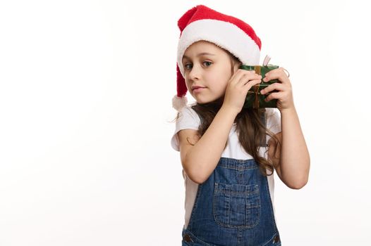 Isolated portrait on white background of a happy, adorable curious little child girl in Santa hat, shaking a Christmas present and guessing what's inside. Copy ad space. Time to open Xmas gift boxes