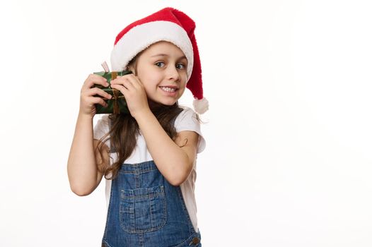 Isolated portrait on white background of a mischievous little girl in Santa hat, shaking a Christmas present and guessing what's inside. Looking forward to the upcoming winter holidays. Copy ad space