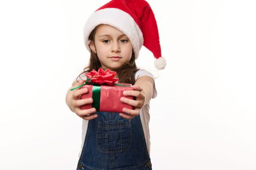 Portrait on white background of a lovely little girl in Santa hat, holding a happy gift box with beautiful tied bow. Christmas Eve. New Year and winter holidays. Time to open presents. Magical mood
