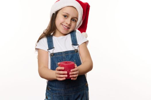 Charming little girl with long hair, wearing Santa hat and blue denim overalls,smiles to camera, posing against a white background with a red decorative Christmas candle in her hands. Copy ad space