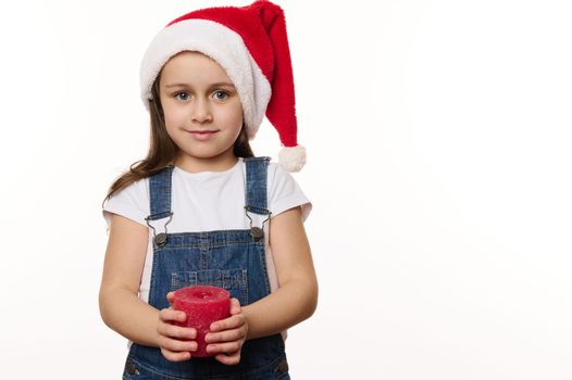 Happy Caucasian little child, lovely baby girl wearing Santa hat and denim overalls, holding a Christmas candle, cutely smiling, looking at camera. Isolated on white background. Copy advertising space