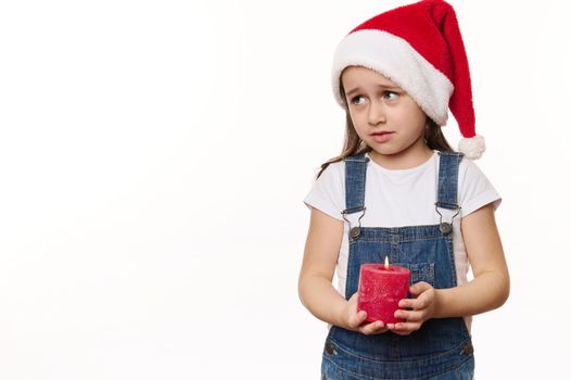 Mischievous little girl in Santa hat, holds a lit candle, makes wish, looks at copy ad space for text on white background. Looking forward to upcoming winter holidays. Merry Christmas. Happy New Year.