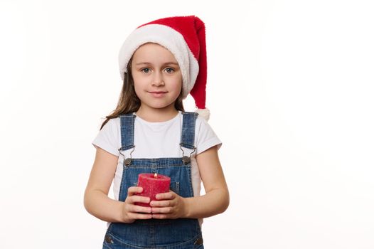 Delightful preschooler girl, lovely little child in Santa hat and blue denim overalls, holds a lit red candle in her hands and cutely smiles looking at camera, isolated on white background. Christmas