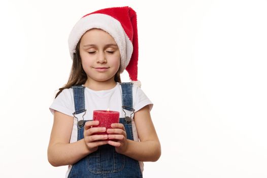 Adorable peaceful little girl in Santa hat, holds a lit candle, makes cherished wish while posing with her eyes closed, against a white background. Copy space. Christmas Eve. New Year. Winter holidays
