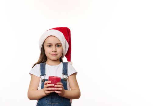 Adorable European baby girl in Santa hat, blue denim overalls, with a lit red candle in hands, isolated over white background. Cute kid makes cherished wish for Christmas. Winter holidays. Copy space