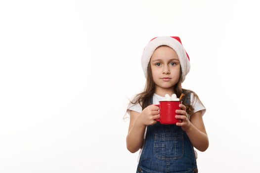 Isolated portrait on white background of a happy little girl in Santa hat, enjoying a delicious sweet hot chocolate drink with marshmallows. Christmas. Winter holidays concept. Copy ad space for text