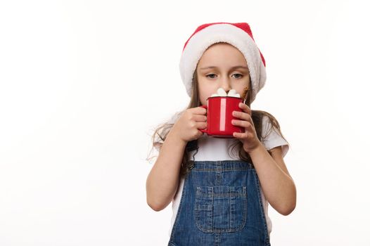 Isolated portrait on white background of a cute baby girl, wearing Santa hat, drinking a delicious hot chocolate with marshmallows. Magical atmosphere of the upcoming Christmas holidays. Celebration