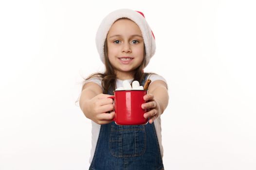 Selective focus on a red cup of hot cocoa drink with marshmallows, in the hands of a blurred lovely baby girl wearing Santa hat. Traditional food and beverage for Christmas. Winter festive atmosphere