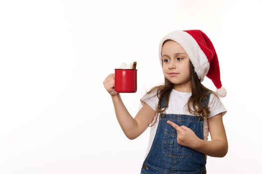 Isolated portrait on white background of a lovely baby girl in Santa, hat pointing at red cup of hot cocoa drink with marshmallows in her hands. Merry Christmas. Winter holiday atmosphere. Copy space