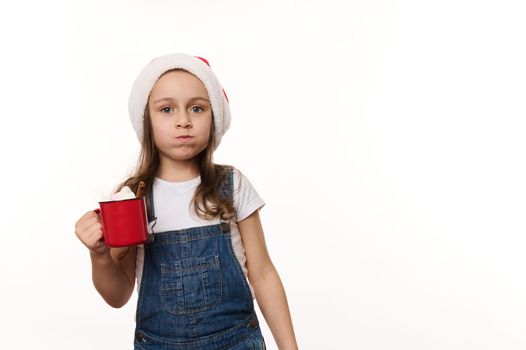 Mischievous 5 years old little girl wearing Santa hat and denim overalls, drinking hot chocolate drink and eating marshmallows, isolated on white background with copy ad space. Christmas atmosphere