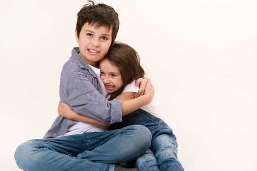 Mischievous Caucasian children, adorable boy and cute girl, brother and his younger sister hugging each other over white background with copy ad space for promotional text. Kids. Friendship concept