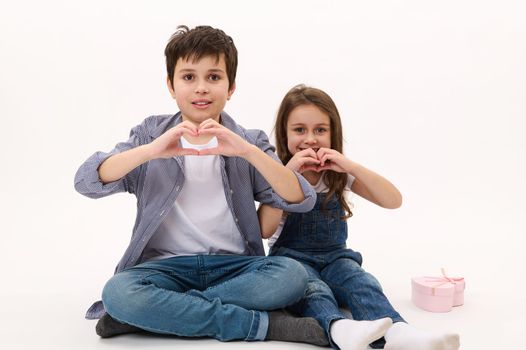 Adorable Caucasian children, boy and girl wearing casual shirt and denim jeans, gesturing showing a heart from fingers, smiling looking at camera, isolated over white background. Saint Valentines Day