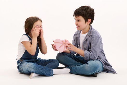 Adorable Hispanic teenage boy, brother gives beautiful heart-shaped pink gift box to his lovely sister, greeting her with St Valentine's Women's Day, isolated on white background. Copy space for text