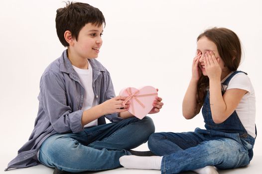 Handsome Caucasian teenage boy, loving caring brother greeting her little sister, who covers her eyes with hands, giving her a cute present in heart-shaped gift box. Saint Valentine's Day. Women's Day