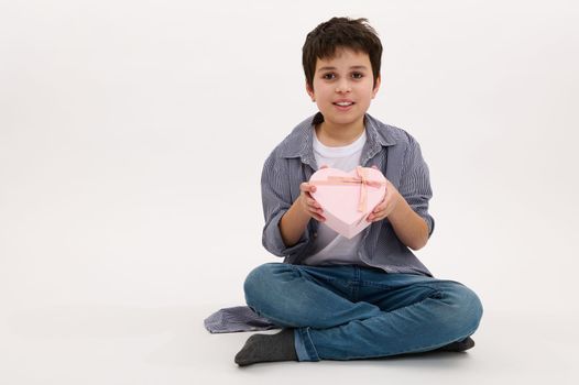 Adorable teenage boy of Middle-Eastern ethnicity holds a beautiful pink heart shaped gift box, isolated on white background with copy space. Saint Valentines Day, Mother's Day. Women's Day