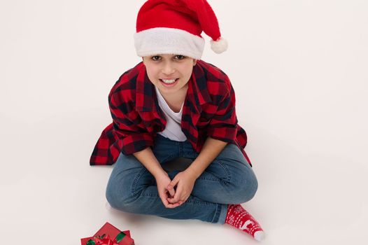 View from above of adorable multi-ethnic preteen boy in Santa hat and red plaid shirt, sitting on white background with christmas presents. Copy advertising space. Isolated portrait. New Year's event