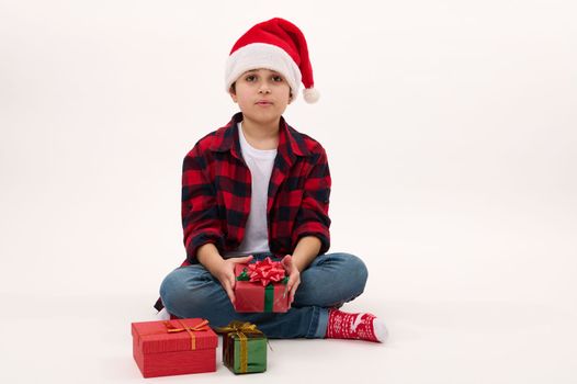 Full length portrait: handsome preteen boy in red plaid shirt and Santa hat, holding a Christmas gift, sitting on white background with copy space for text. Looking forward to upcoming winter holidays