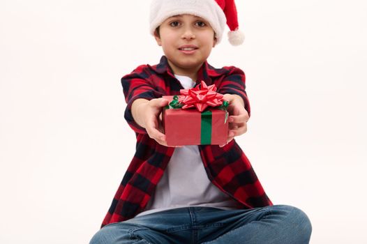 Details: cute Christmas gift with red bow in the hands of a blurred boy wearing Santa hat, hands a present, looking at camera, isolated over white background with copy ad space. New Year. Boxing Day.